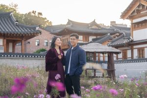 Two people walk through a traditional Korean hanok village with pink flowers blooming in the foreground.