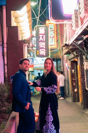 A couple walks through a colorful nighttime street with Korean neon signs.