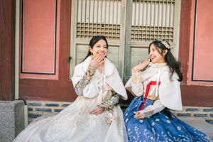 Two people in colorful traditional Korean hanbok dresses posing against a red wall.