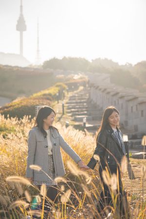 Two friends walking through tall golden grass at sunset with houses and a tower in the background.