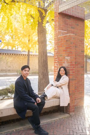 A couple sits together on a brick ledge surrounded by golden autumn trees and dappled sunlight.