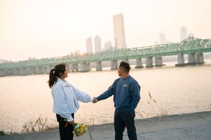 People walking along waterfront with city skyline and bridge in the distance at sunset.