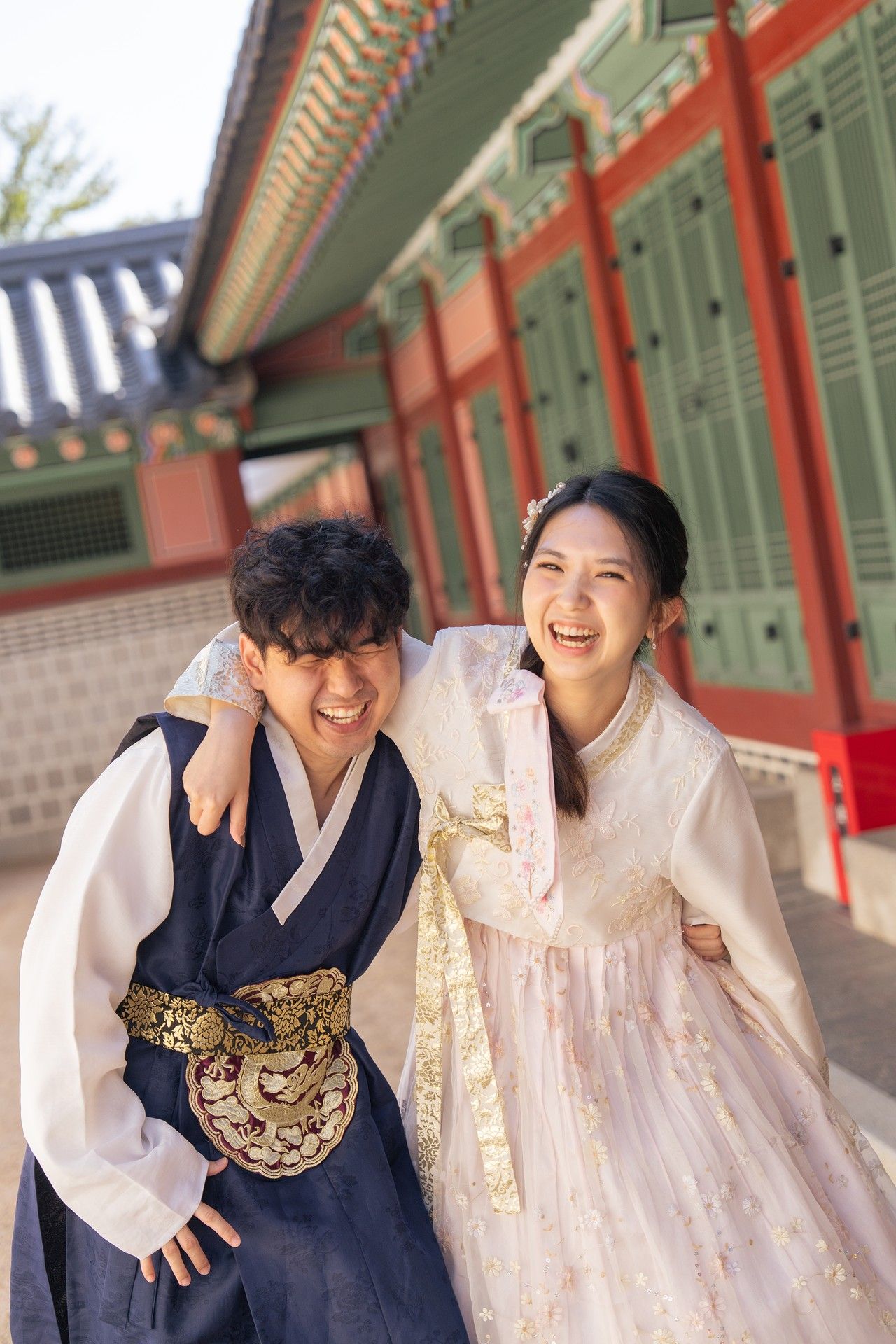 Two people in traditional Korean hanbok clothing share a joyful moment at a historic palace building.