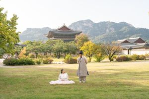 A couple walks together in traditional Korean hanbok attire through a scenic mountain landscape at sunset.
