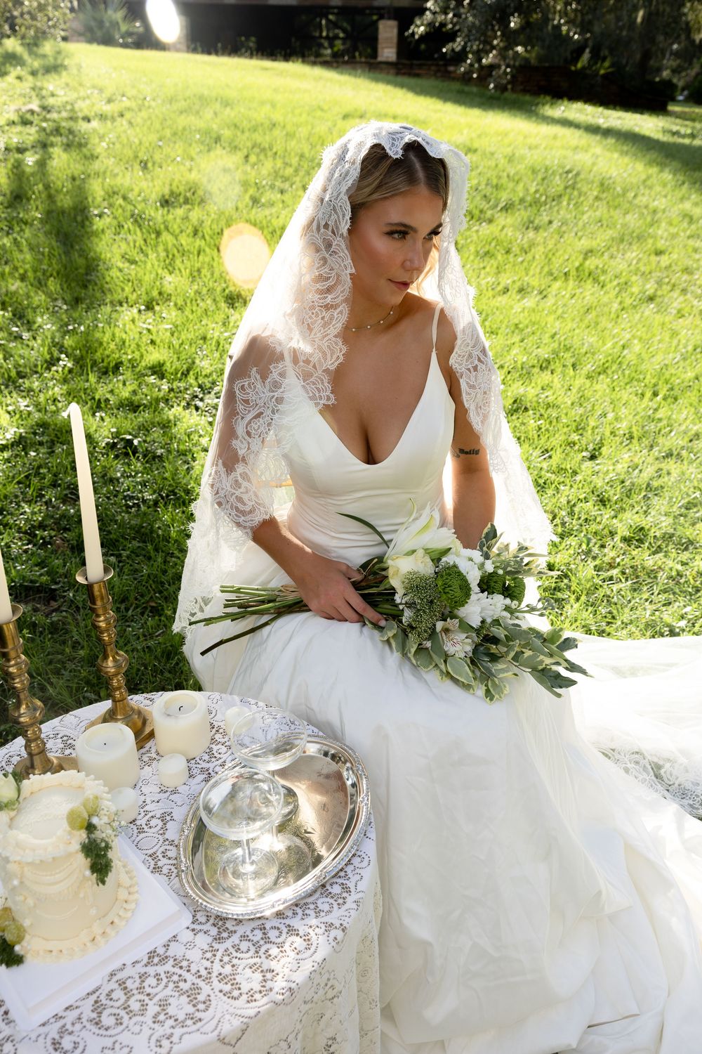 A romantic outdoor wedding table setting with white lace tablecloth, candles, and floral arrangements in a garden.
