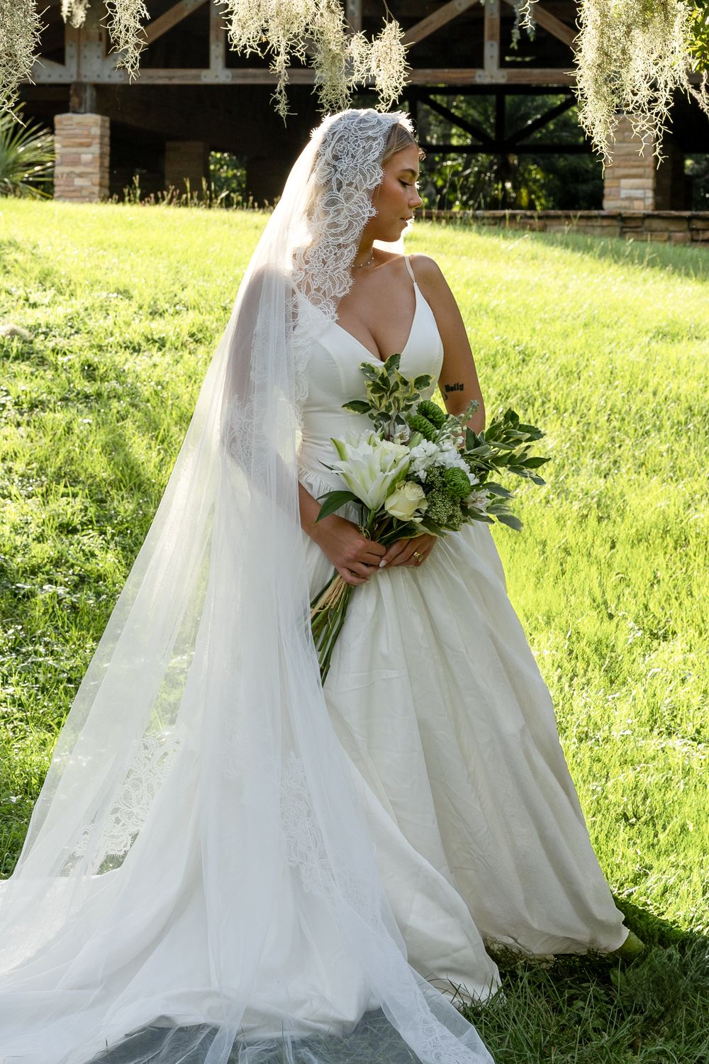A classic white wedding gown with lace veil flows elegantly on a sunny grassy lawn surrounded by greenery.
