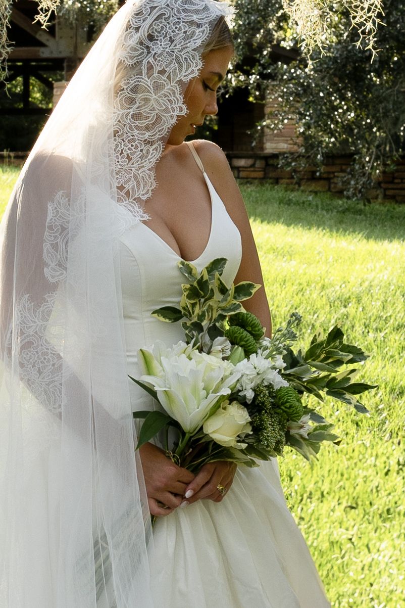 A beautiful bridal portrait showing an elegant lace veil and white lily bouquet against a sunlit garden backdrop.