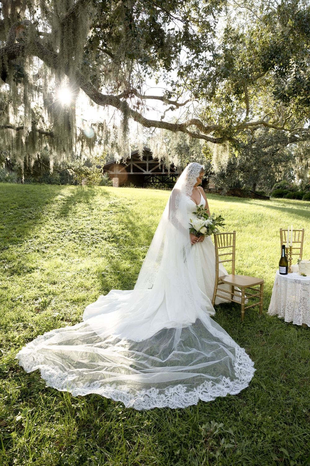 A bride in a flowing white lace gown with a long train sits gracefully on a wooden chair in a sunlit garden setting.