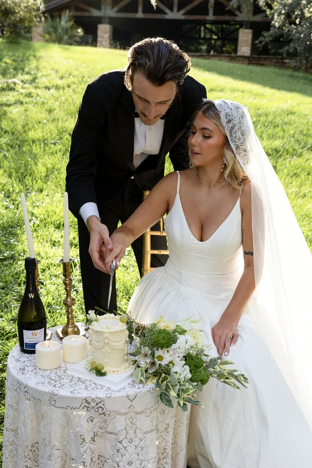 Elegant wedding celebration scene with champagne bottles and white decorations on a lace-covered table outdoors.