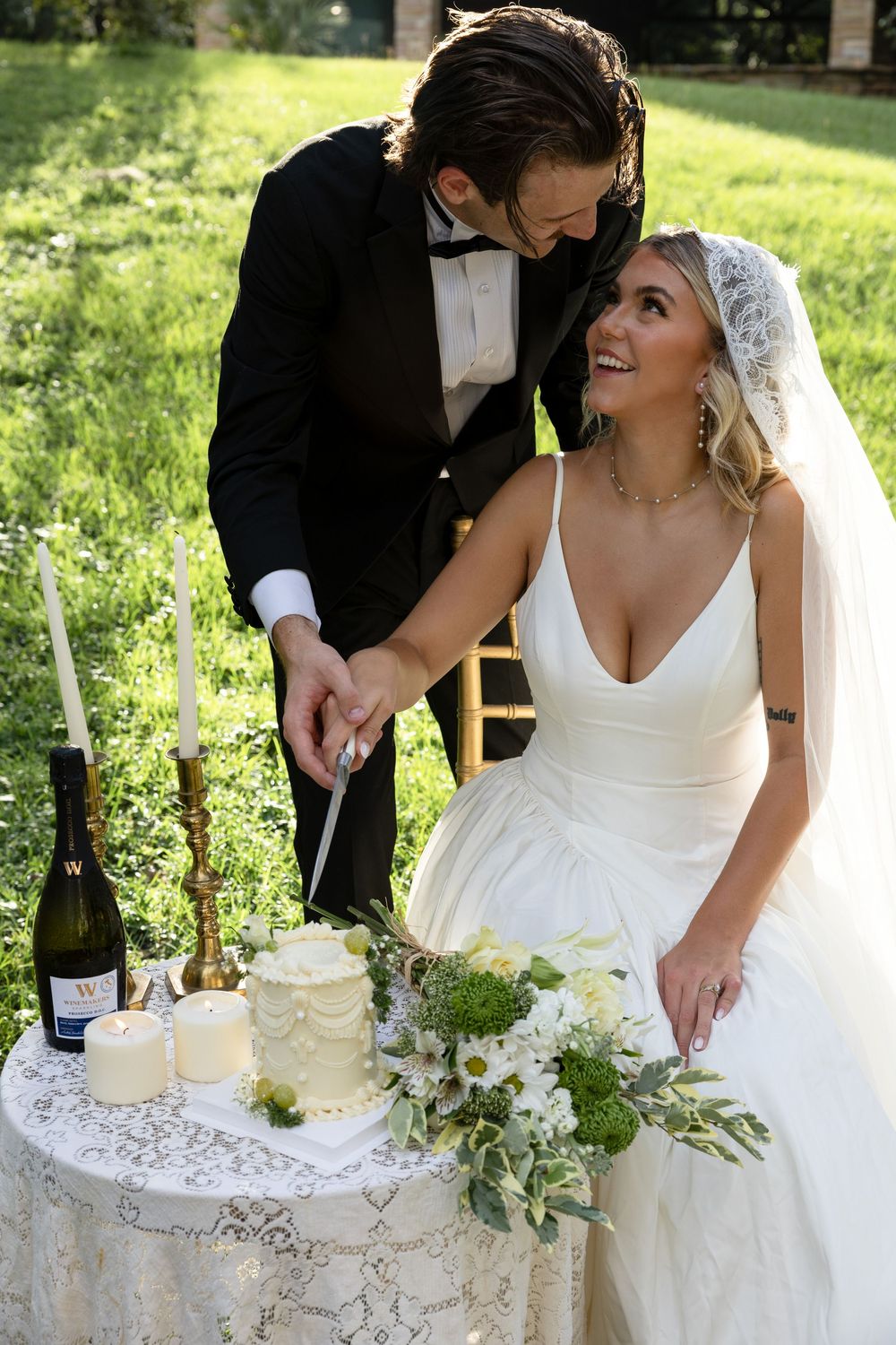 A wedding couple cutting their white tiered cake together on a lace-covered table outdoors in a garden setting.