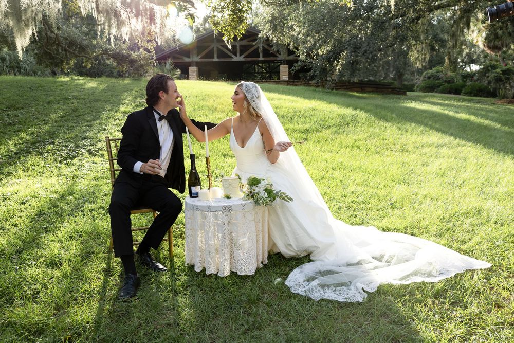 A newlywed couple sharing a special moment while sitting at their outdoor reception table with a cake.