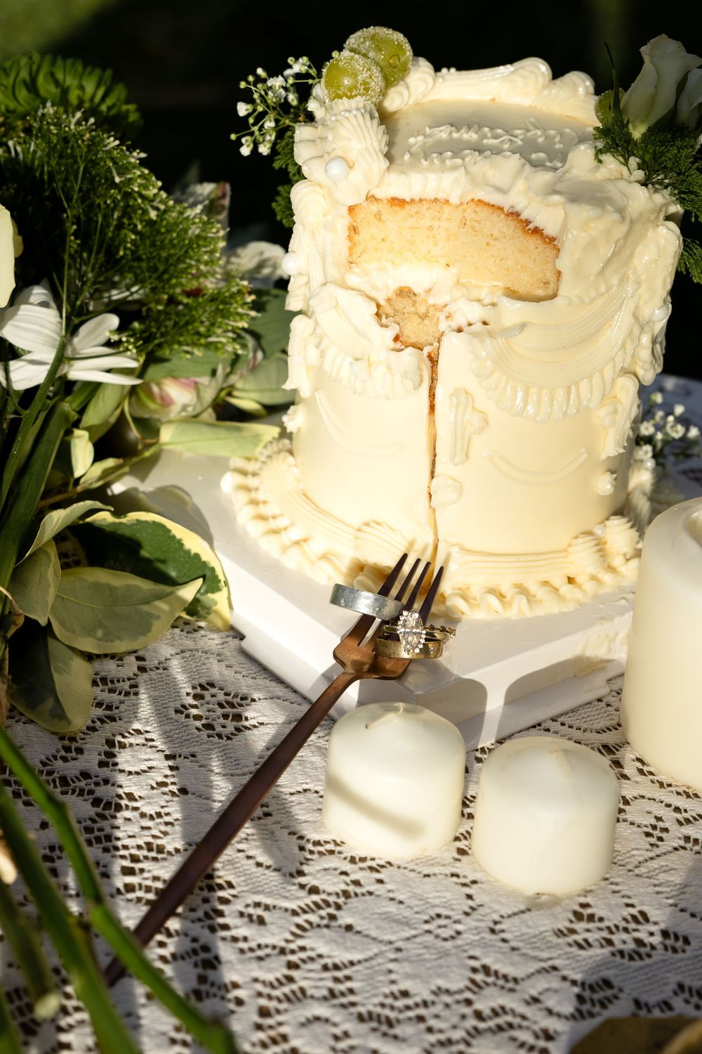 Sliced white wedding cake with delicate frosting details sits on lace tablecloth surrounded by greenery and white floral decor.