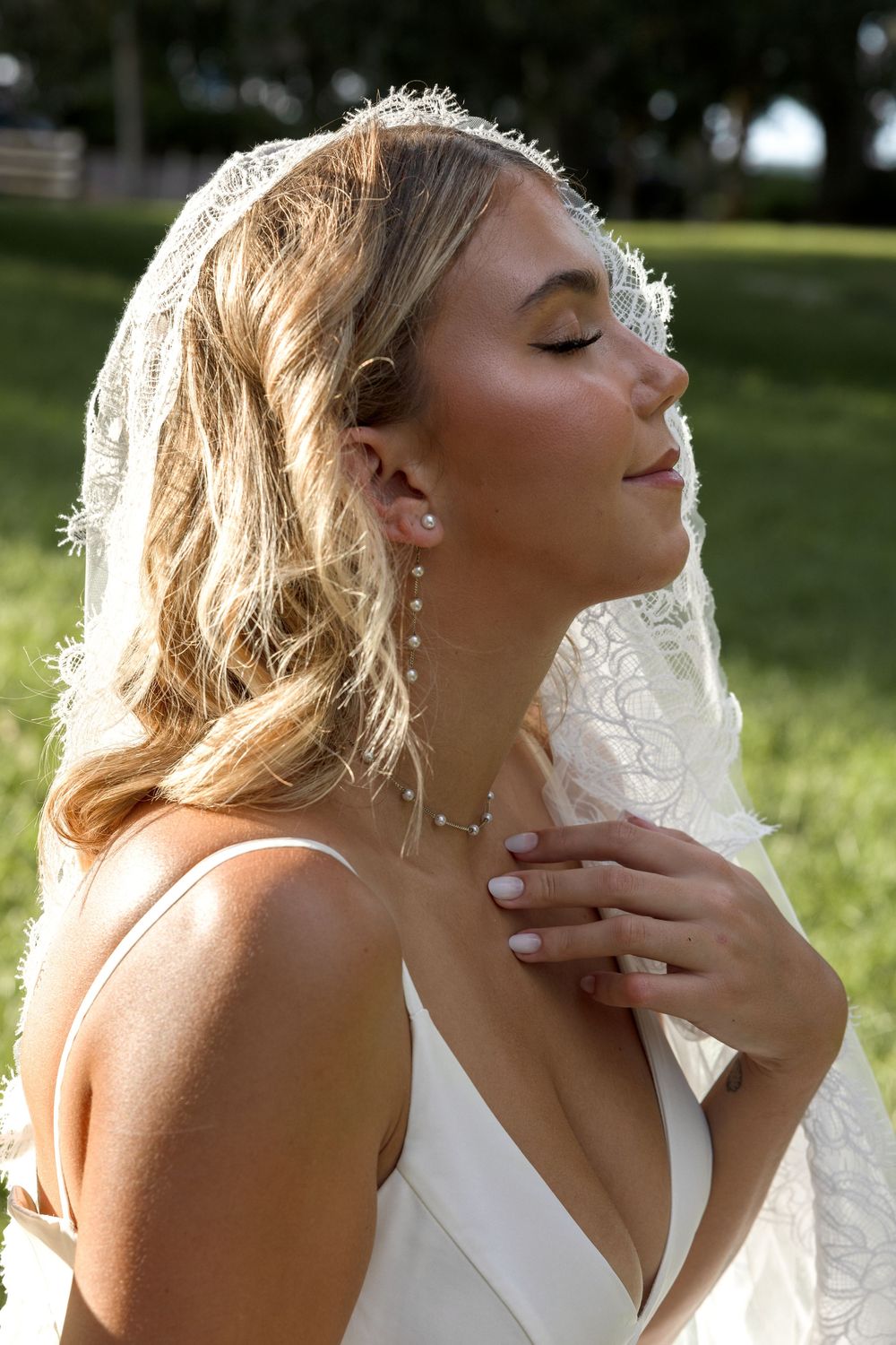 A bride in a white wedding dress tilts her head back in sunlit outdoor setting wearing delicate jewelry.