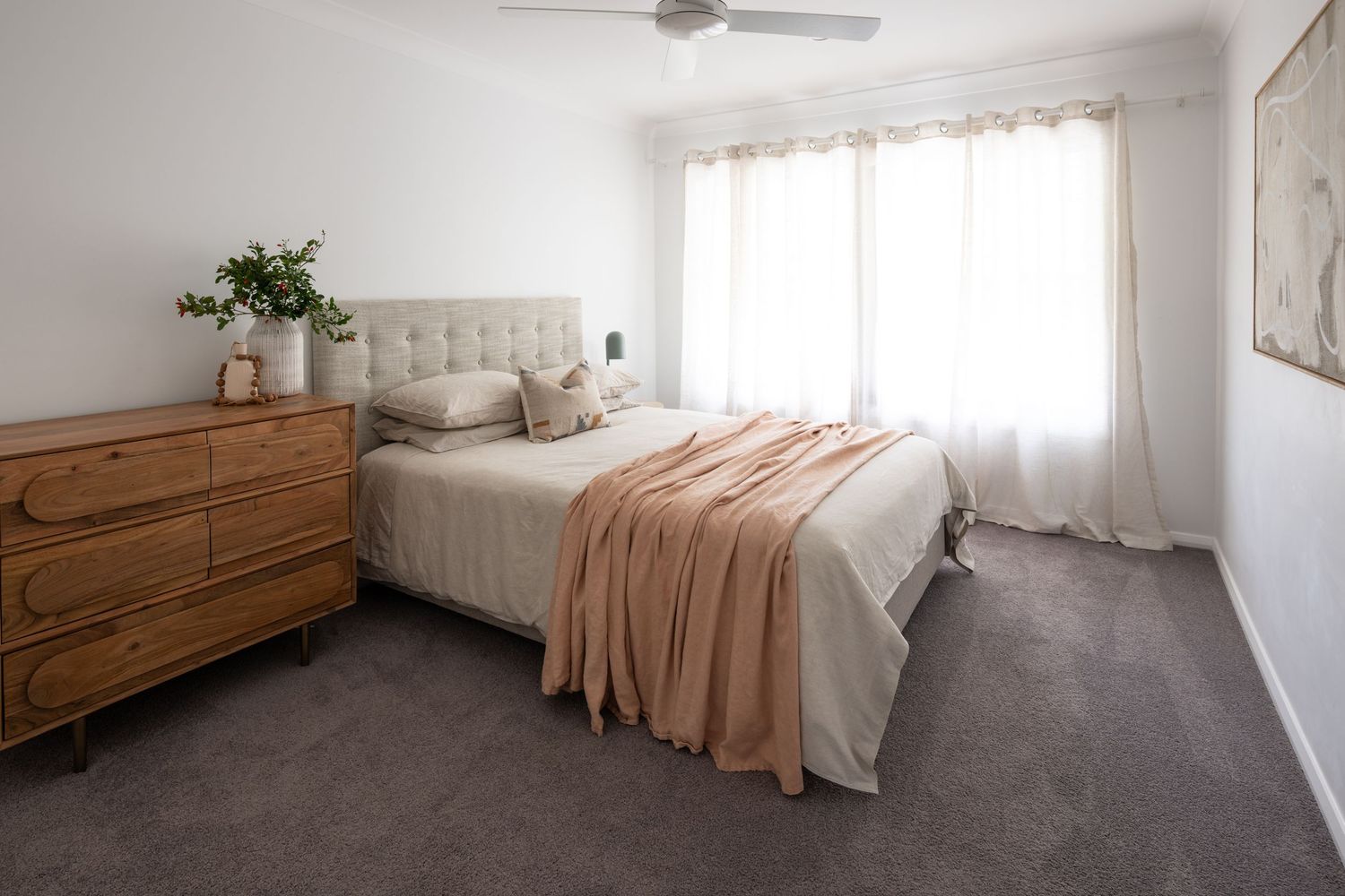 Minimalist bedroom interior with wooden furniture, beige bedding, and ceiling fan in natural lighting.