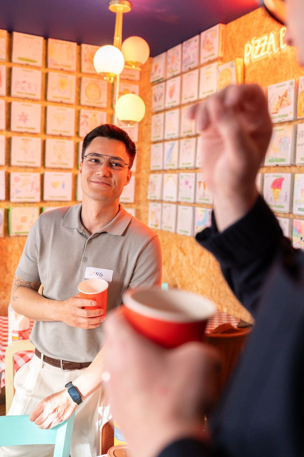 A person in a grey polo shirt holds a red cup while someone gestures in the foreground during a business gathering in Cambridge