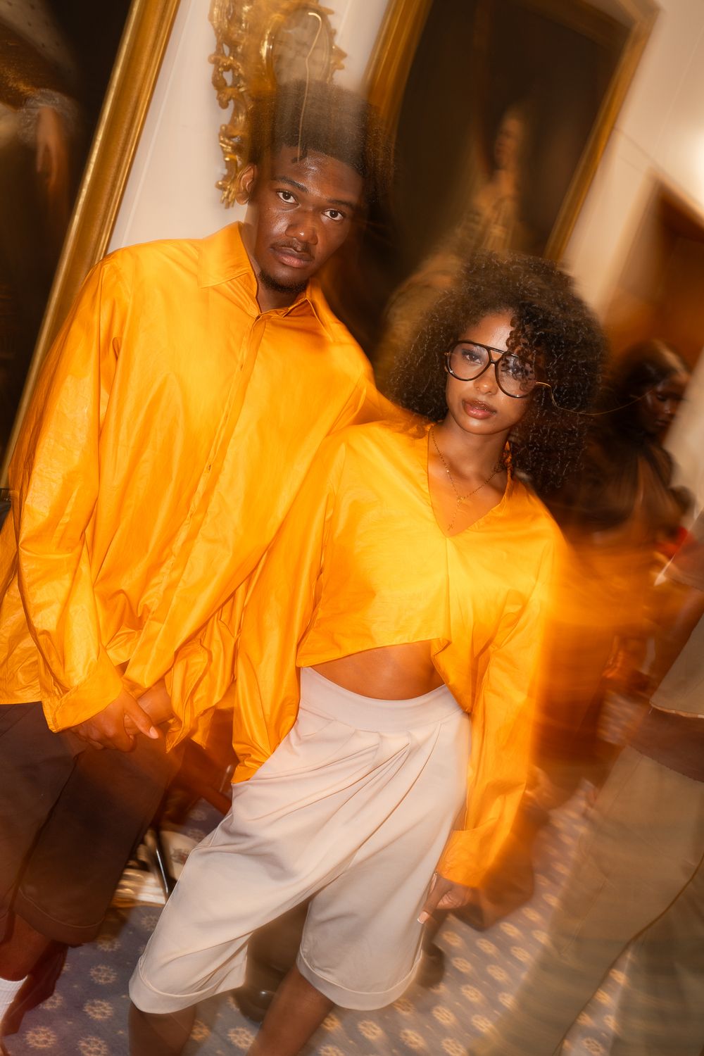 Two models wearing matching bright orange cropped tops and white bottoms pose together indoors at Jimmy Choo fashion show in London