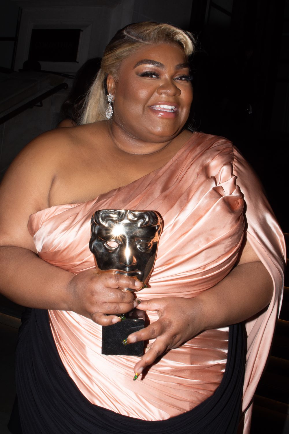 Davine Joy in a peach-colored dress holds a BAFTA award at an evening BAFTA ceremony in London