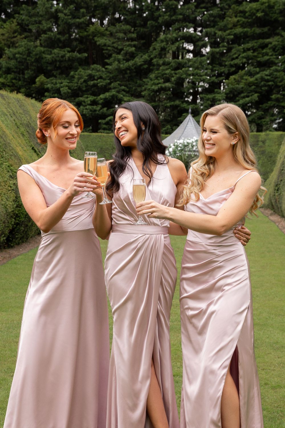 Three women in blush pink bridesmaid dresses laugh together during an outdoor wedding photo session at Swynford Manor, Cambridge