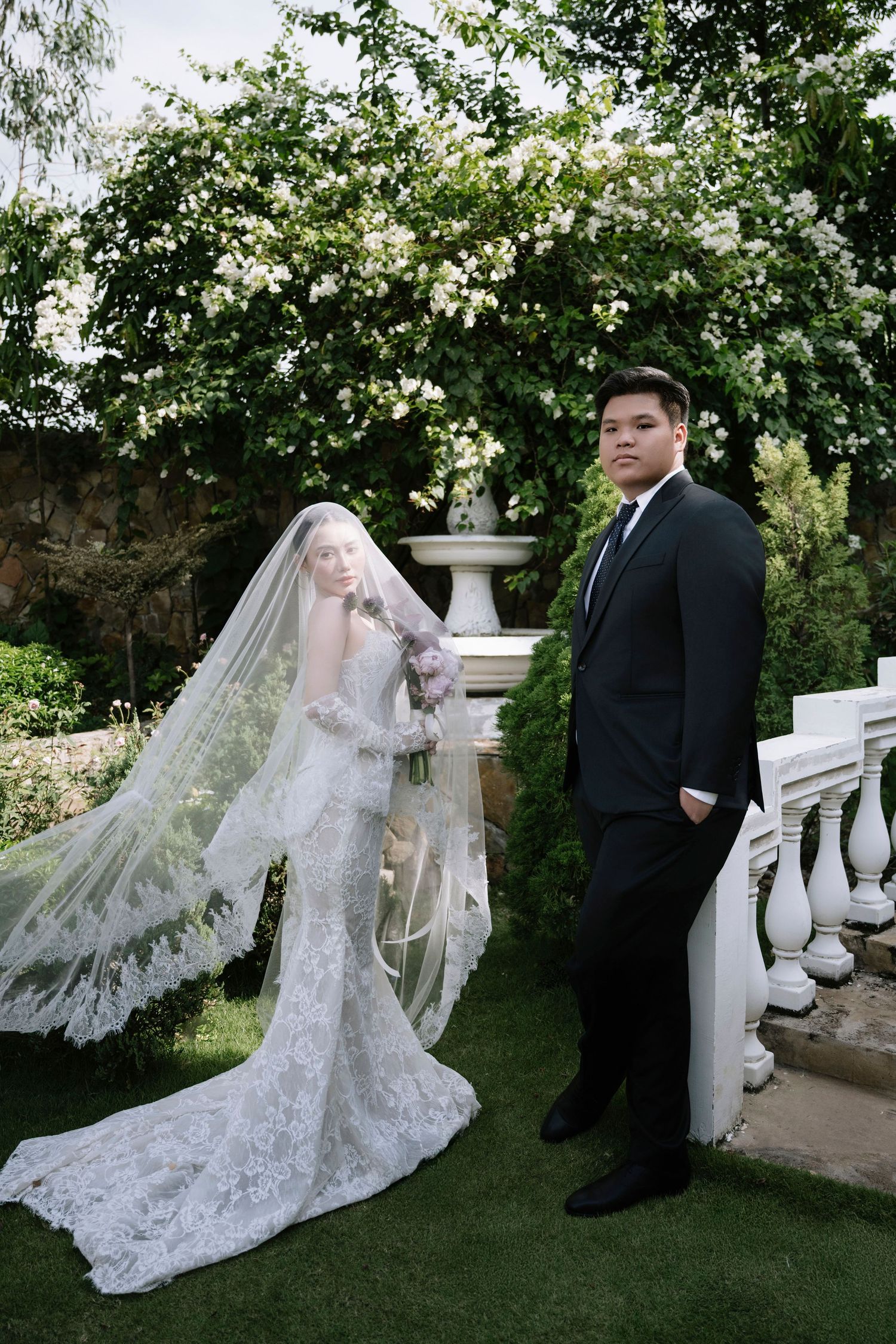 Wedding couple posing in a lush garden, bride in long lace gown with veil, groom in black suit near white roses.