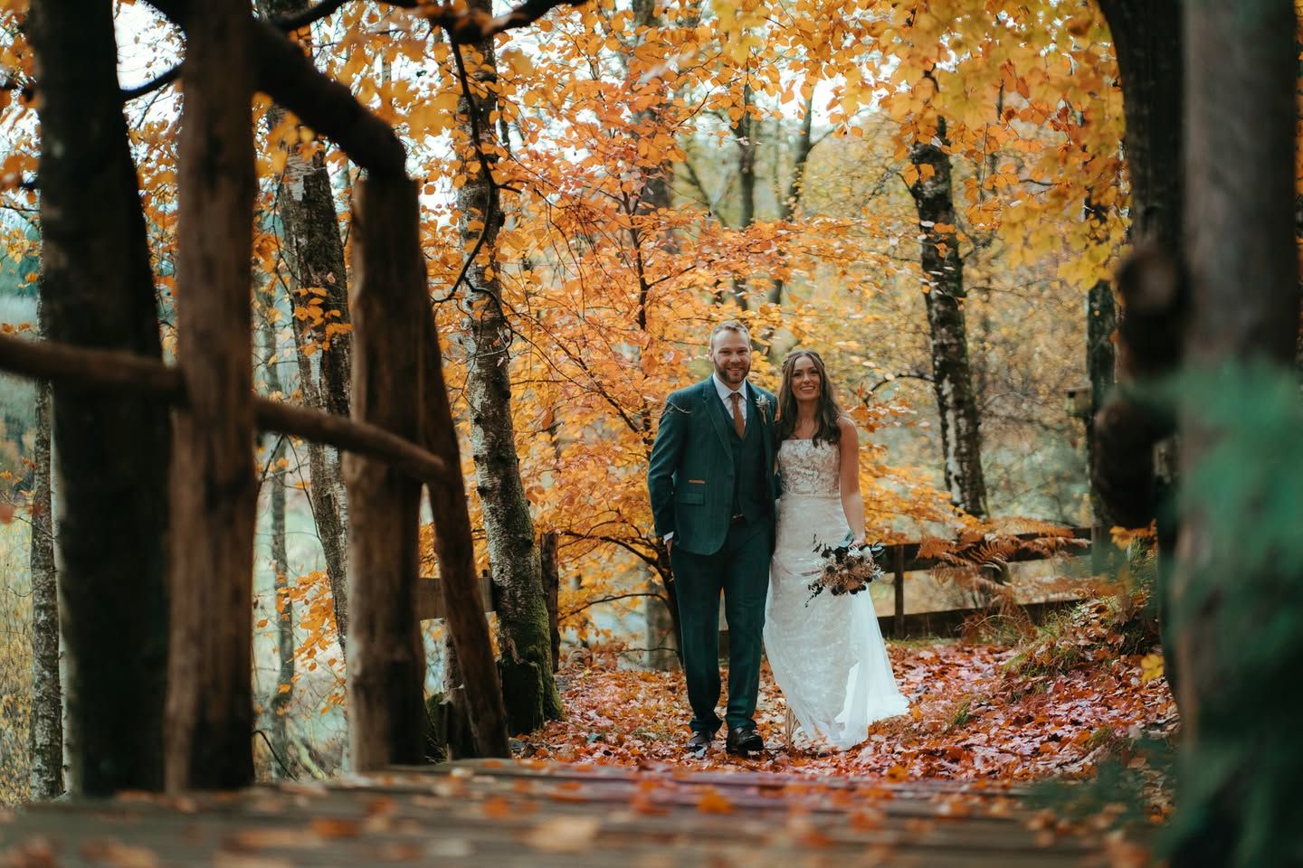 A bride and groom walk hand in hand through a stunning autumn forest with vibrant orange foliage.