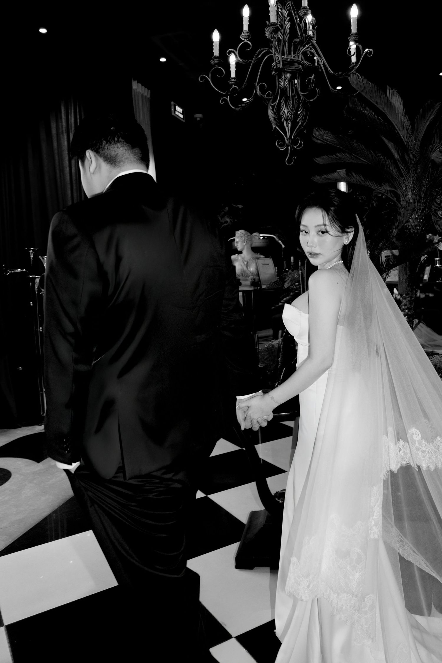 Black and white photo of bride and groom holding hands on checkered floor under chandelier.