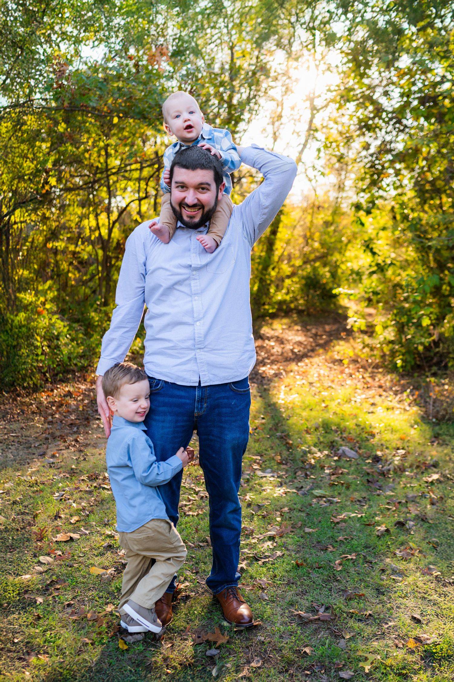 Dad holds infant son on shoulders while preschool son hugs his leg during outdoor fall family photos in richardson.