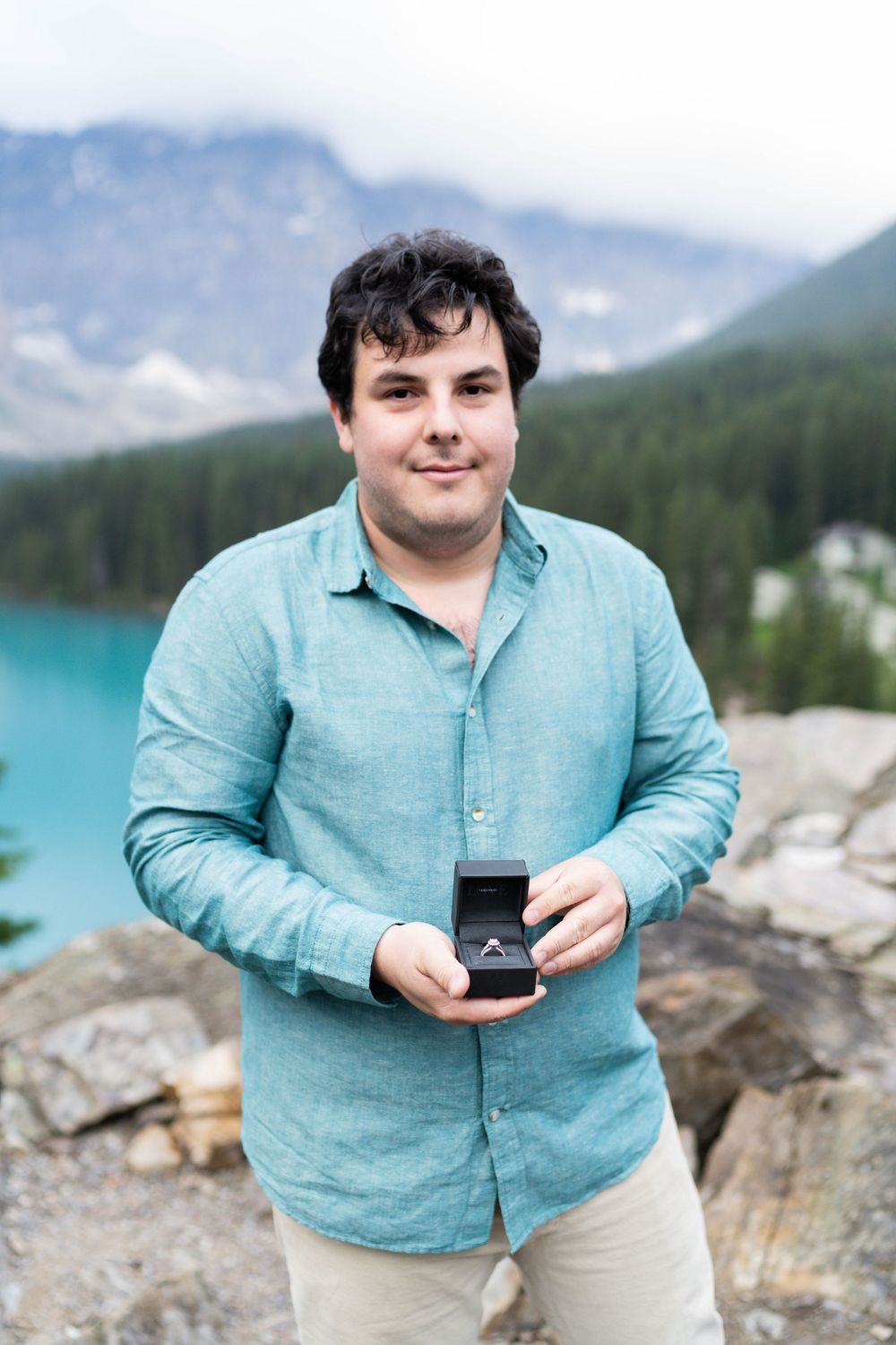 Person in turquoise shirt holding engagement ring box against mountain backdrop at Lake Moraine.