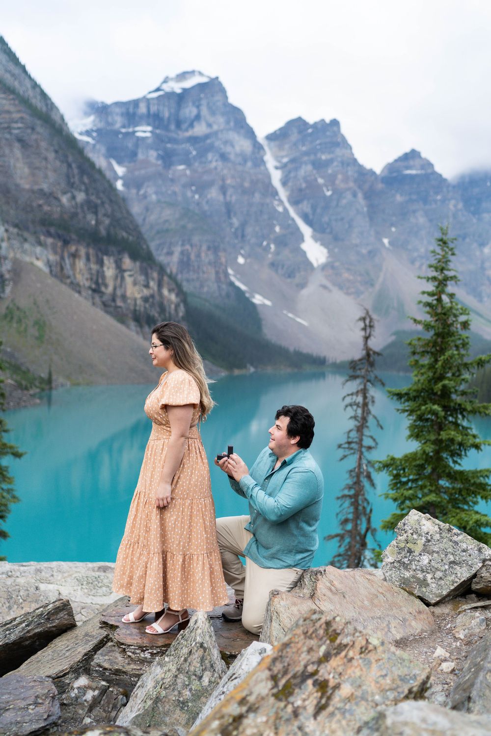 A couple shares a romantic proposal moment against the stunning backdrop of turquoise Moraine Lake and Rocky Mountains.