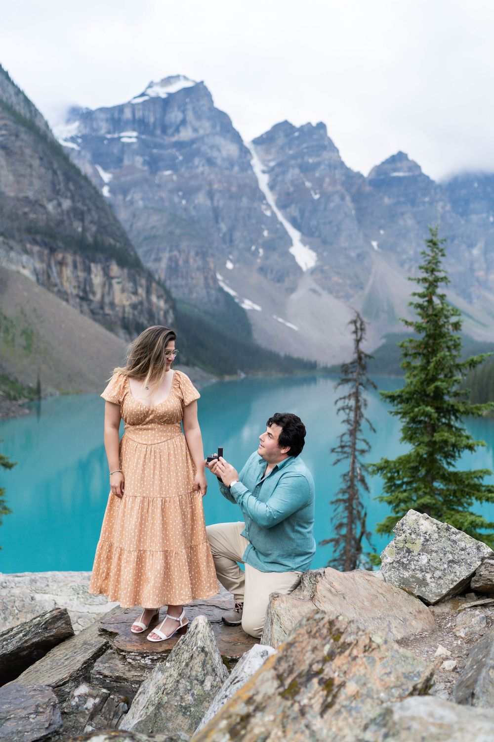Couple sitting on rocky overlook with turquoise Lake Moraine and mountain peaks in background.