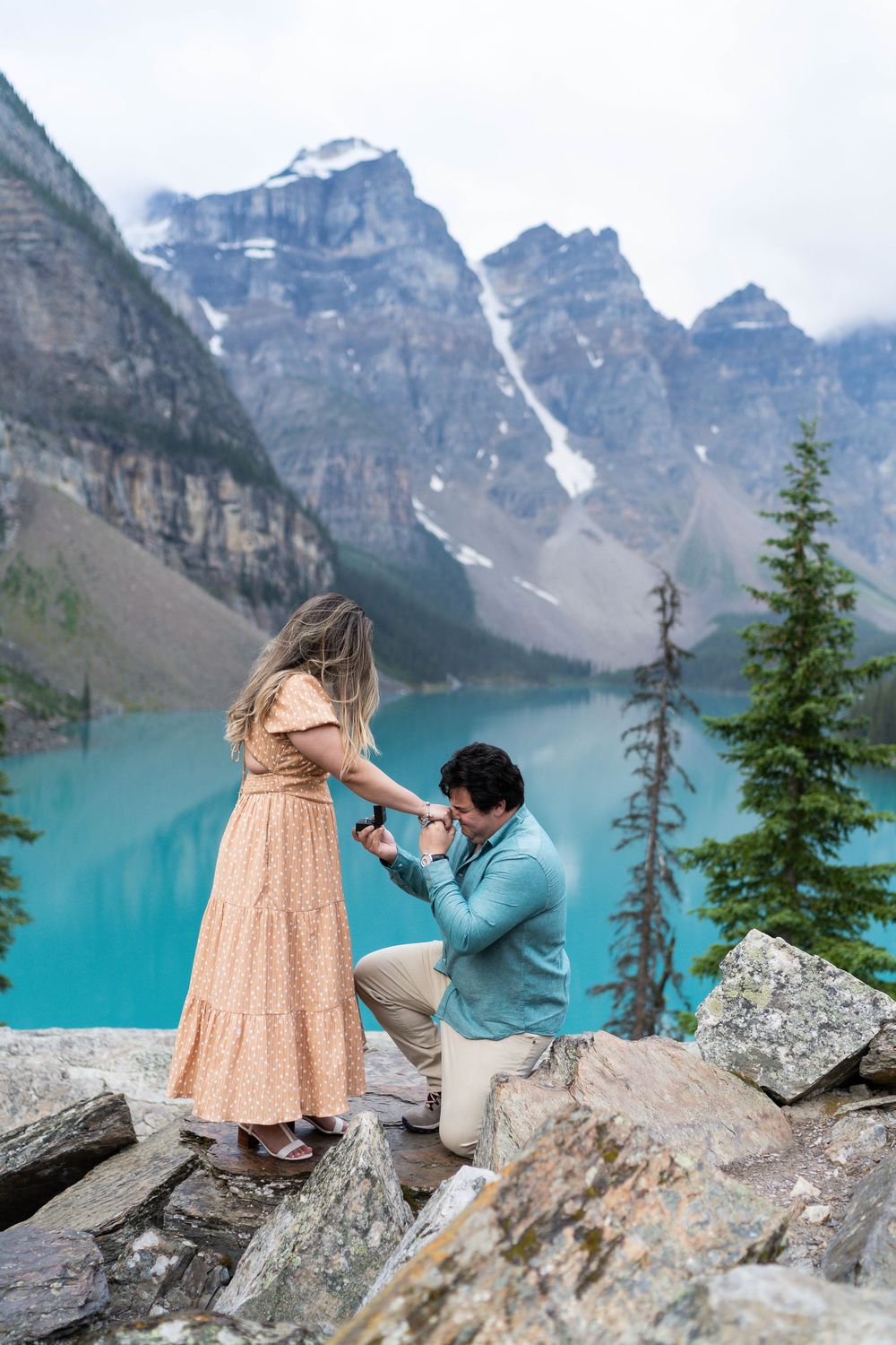 A romantic proposal moment with a stunning turquoise alpine lake and snow-capped mountain peaks in the background.