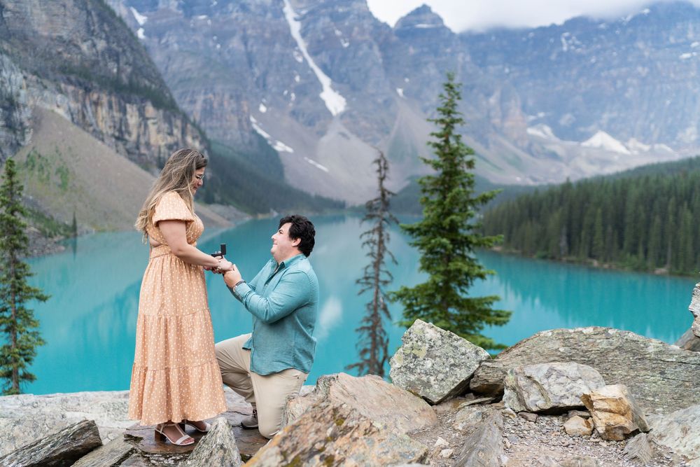 Couple embracing on rocky overlook with stunning mountain peaks and turquoise waters below.