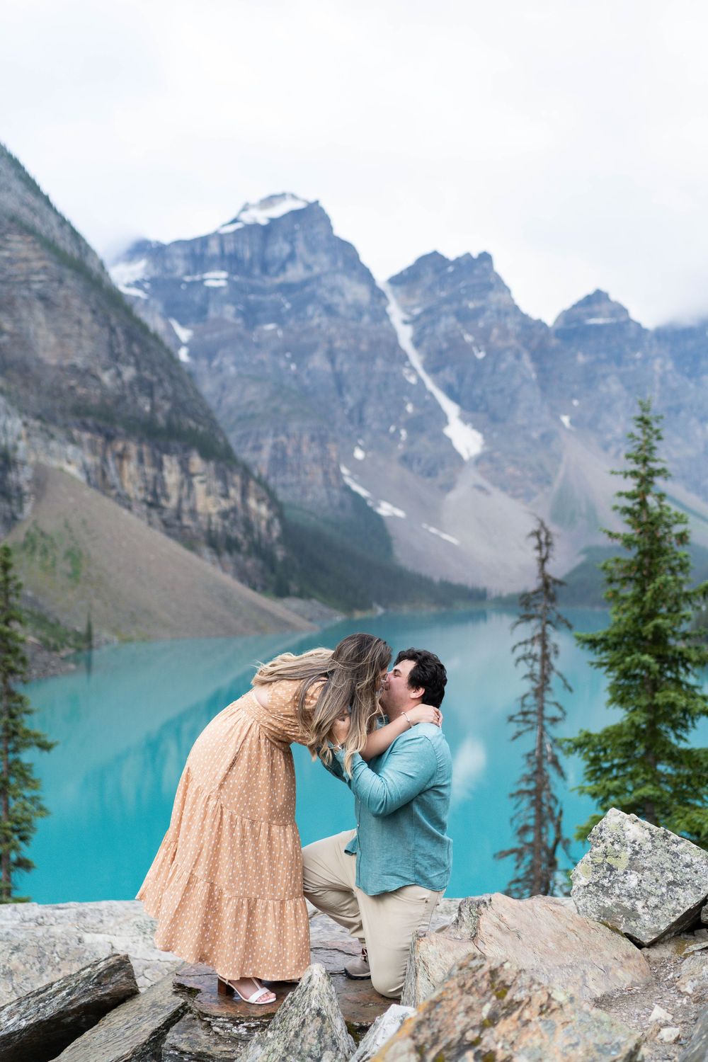 A couple share a romantic embrace at Moraine Lake in Banff National Park with turquoise waters and mountain peaks behind them.