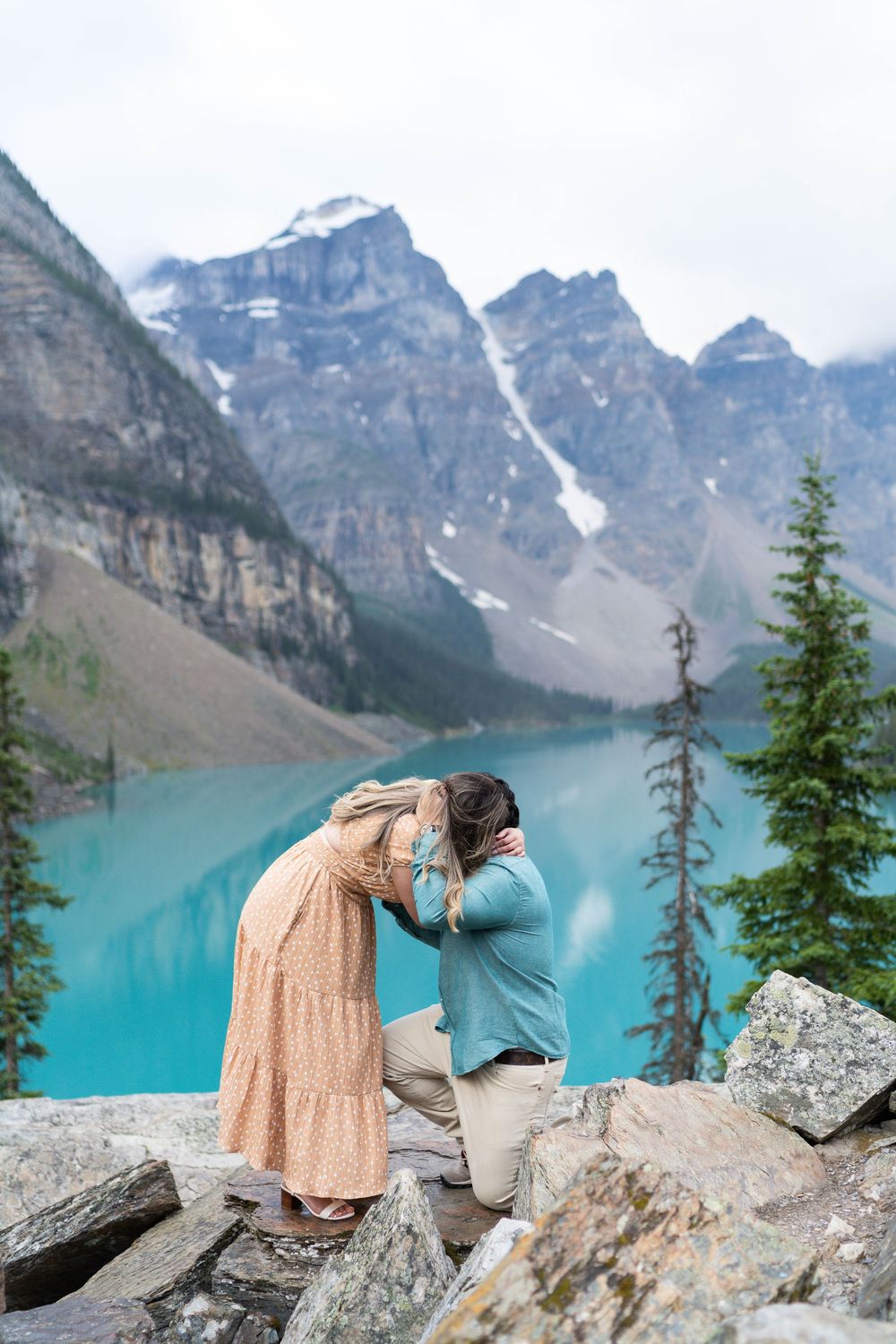 Person in peach dress poses dramatically on rocky ledge overlooking turquoise alpine lake and snow-capped mountains.