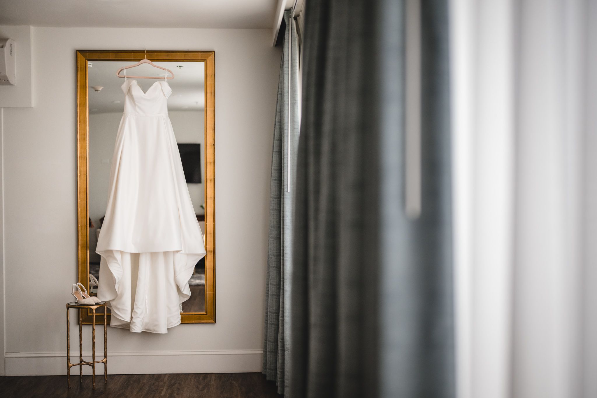 Elegant white wedding dress hanging in front of a gold-framed mirror with natural window light.