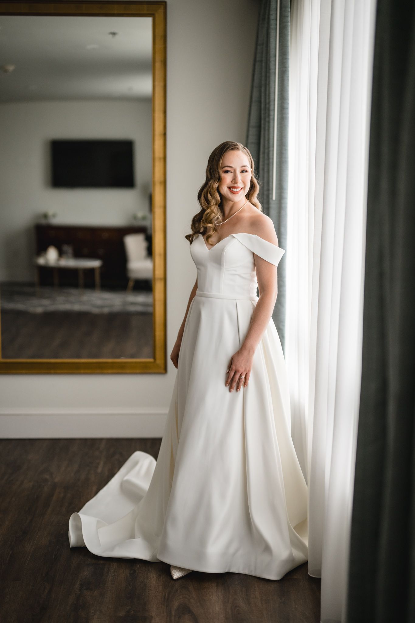 A stunning white wedding dress with an off-shoulder neckline photographed in a bright room with natural lighting.