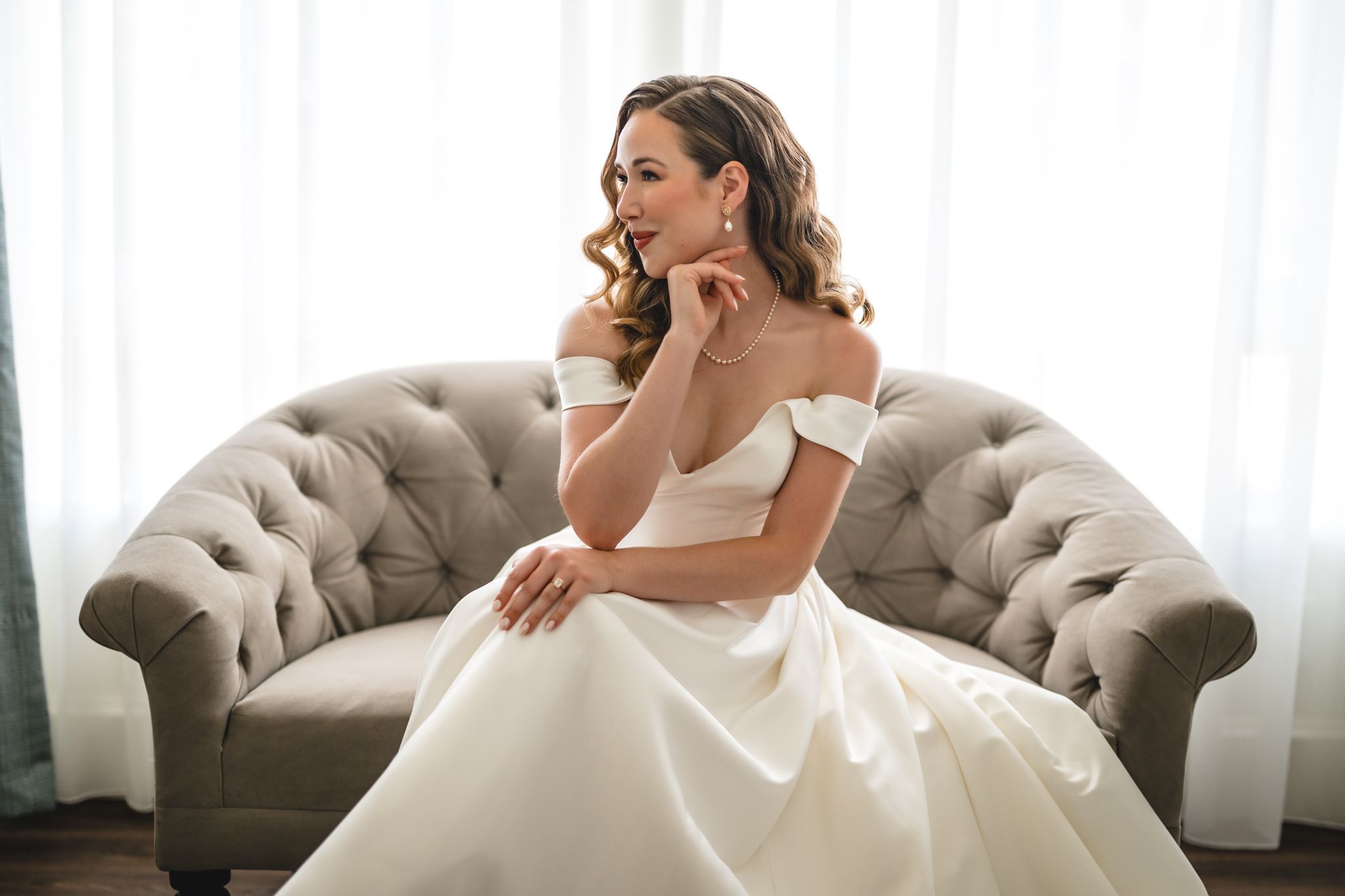 A bride in a luxurious white ball gown poses on a tufted grey couch near a window.