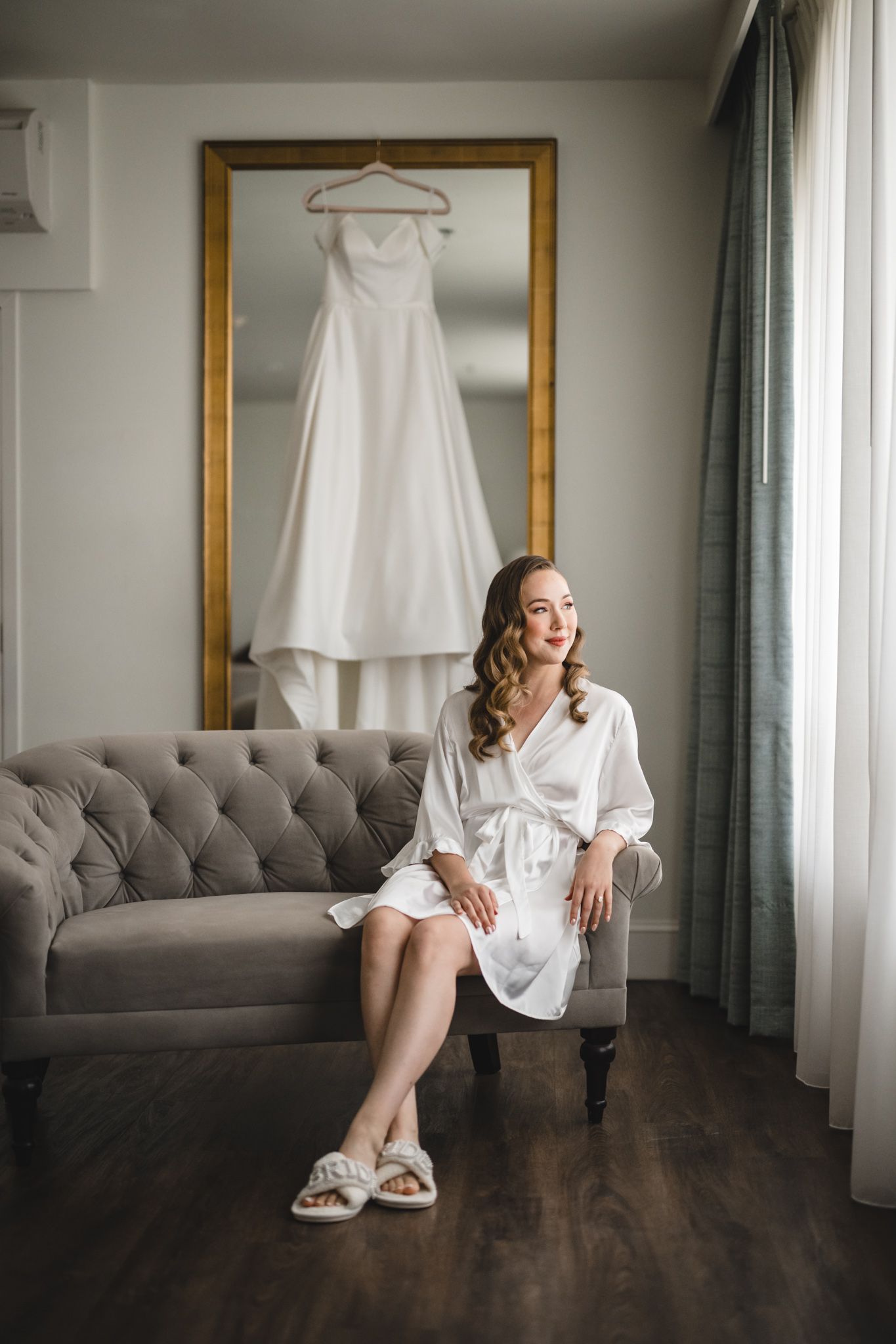 Wedding preparation scene with white dress hanging and grey tufted sofa in elegant room.