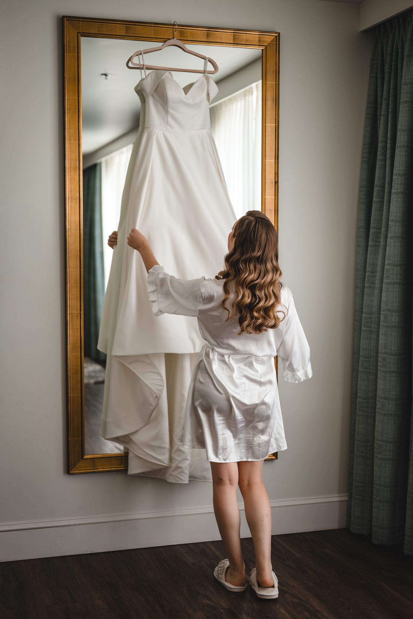 White wedding dress displayed on hanger against gold-framed mirror in sunlit room.