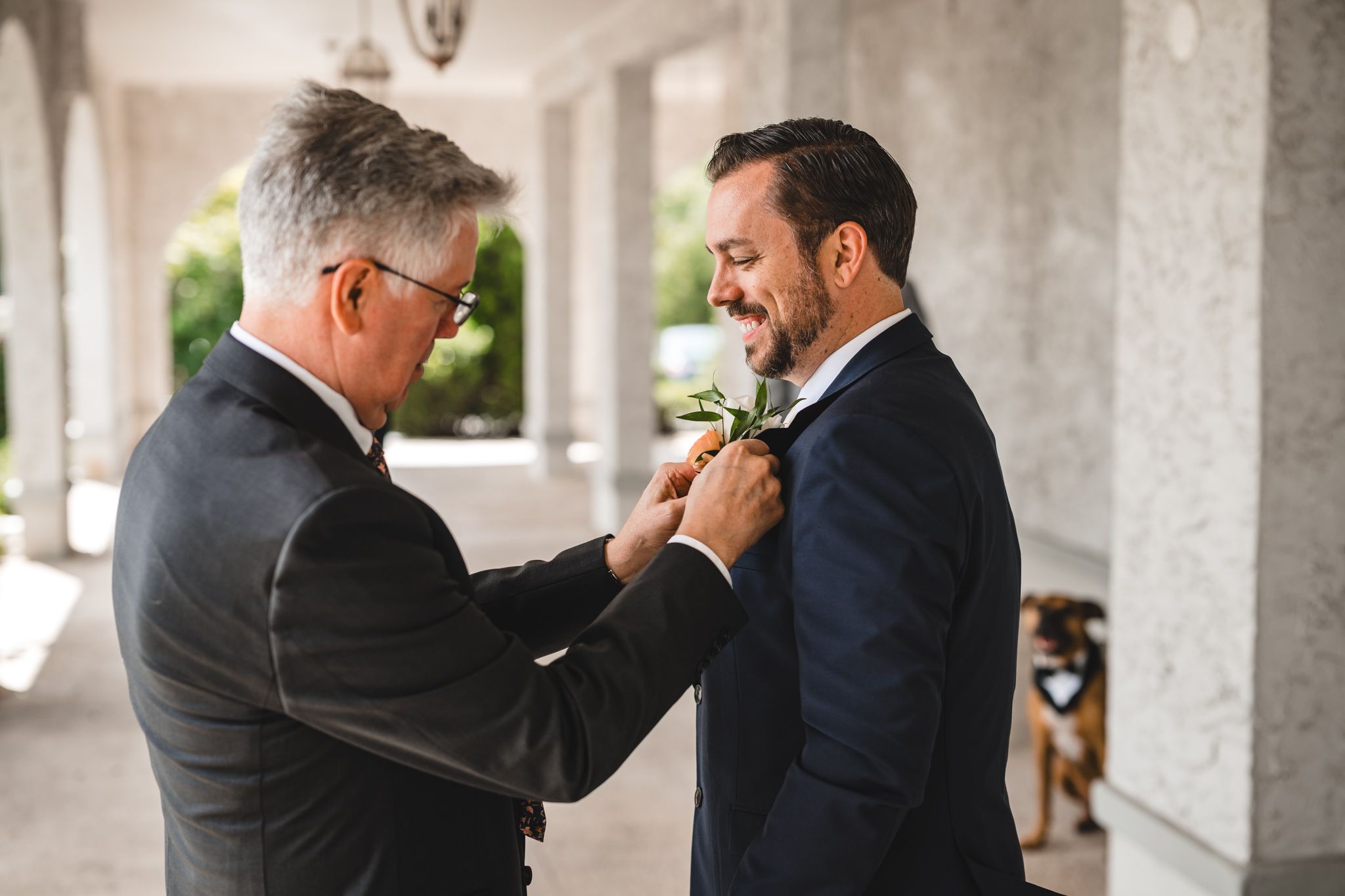 Two men in formal attire share an emotional moment in a columned outdoor setting.