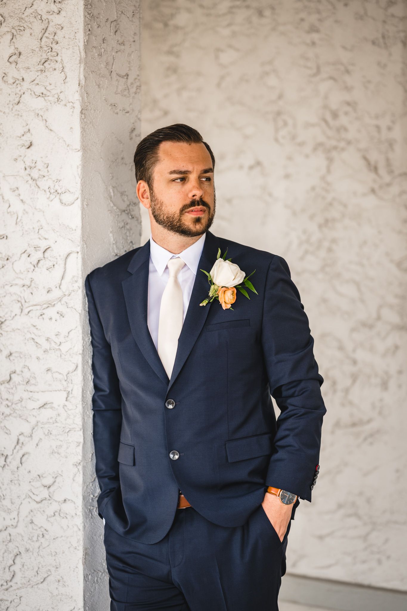 A groom in a navy suit and white boutonniere poses against a textured wall.