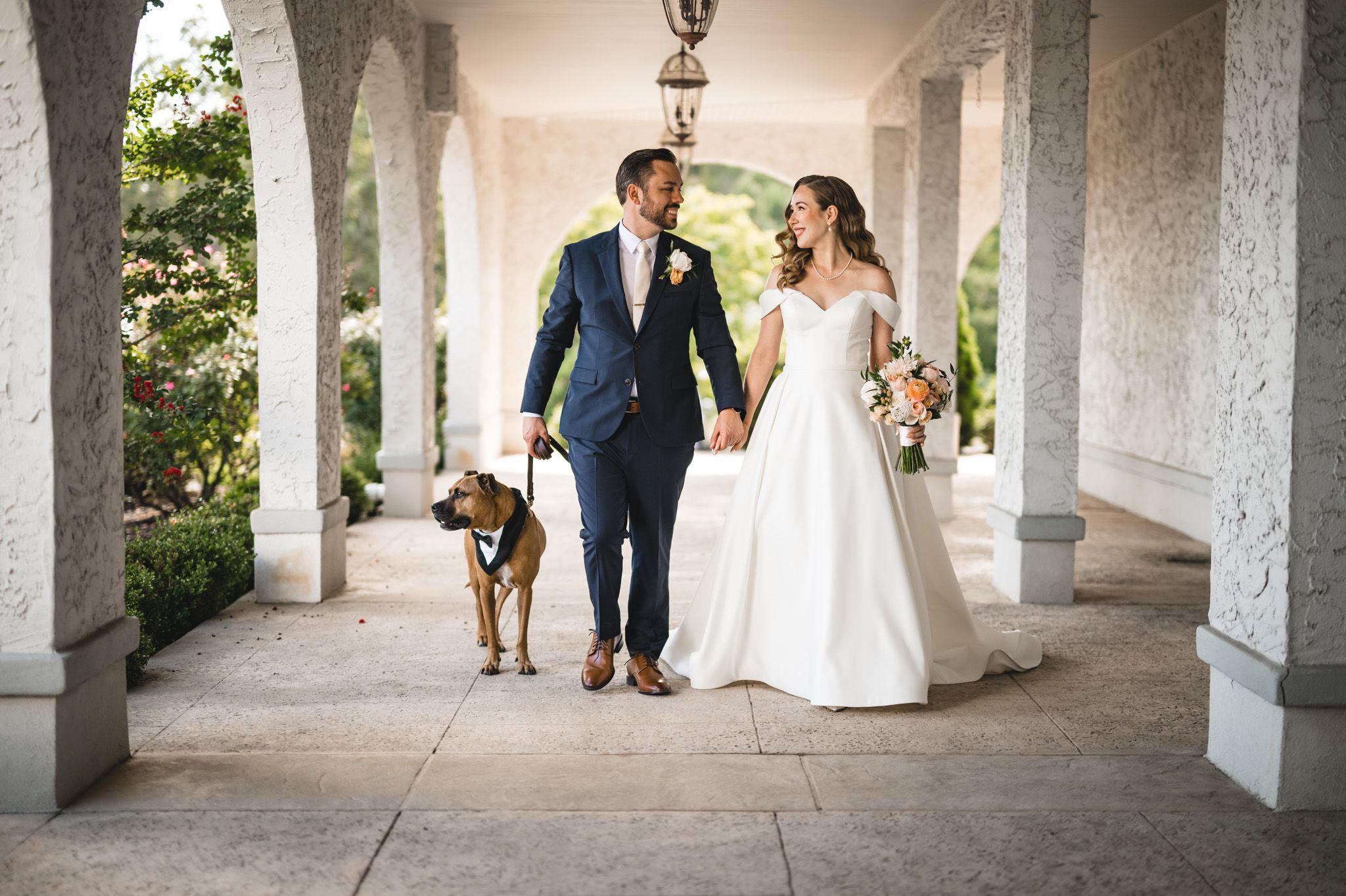 Wedding couple poses under outdoor colonnade with white pillars and natural light.