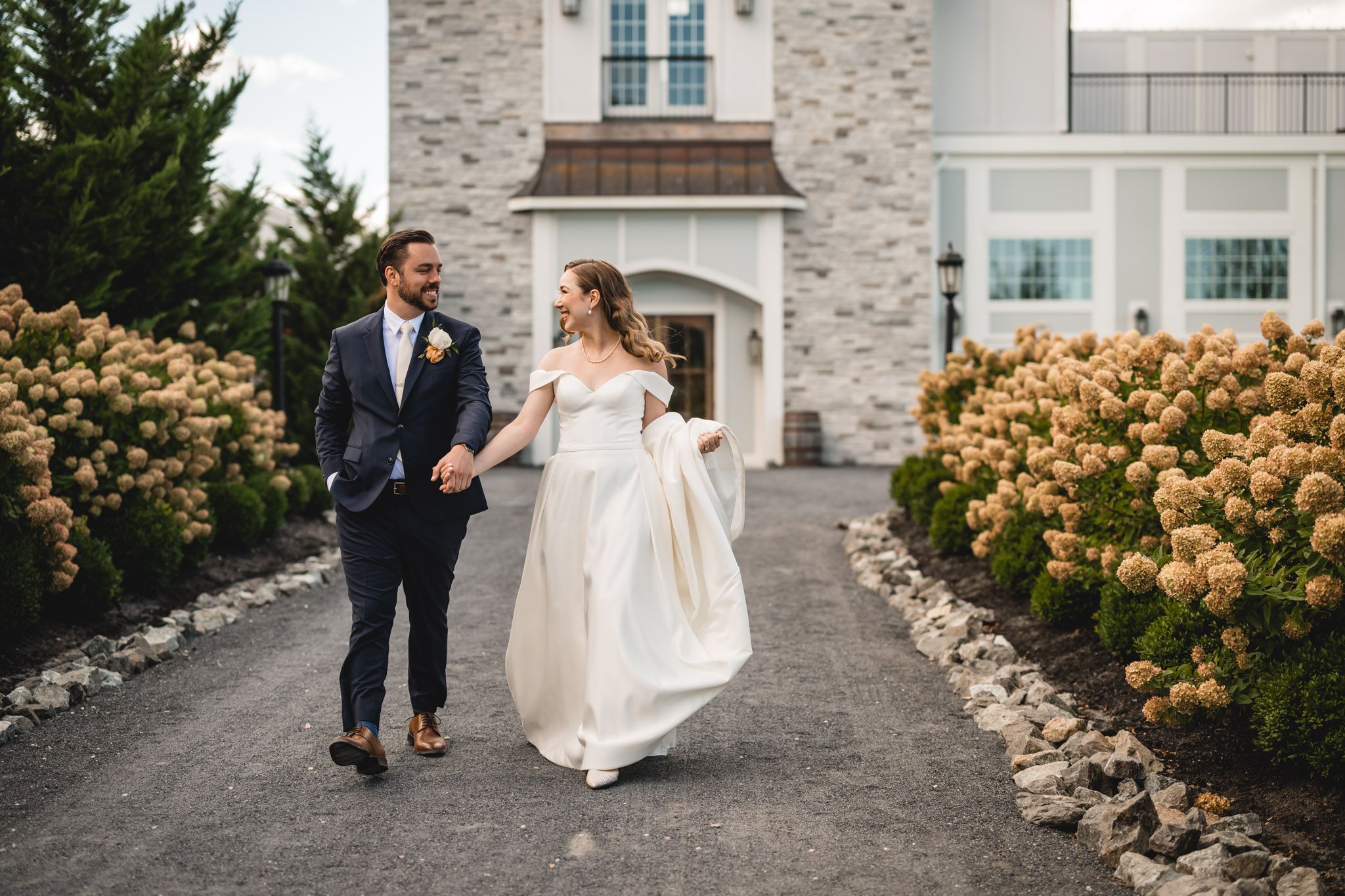 A couple walks along a path lined with blooming flowers outside a stone mansion.