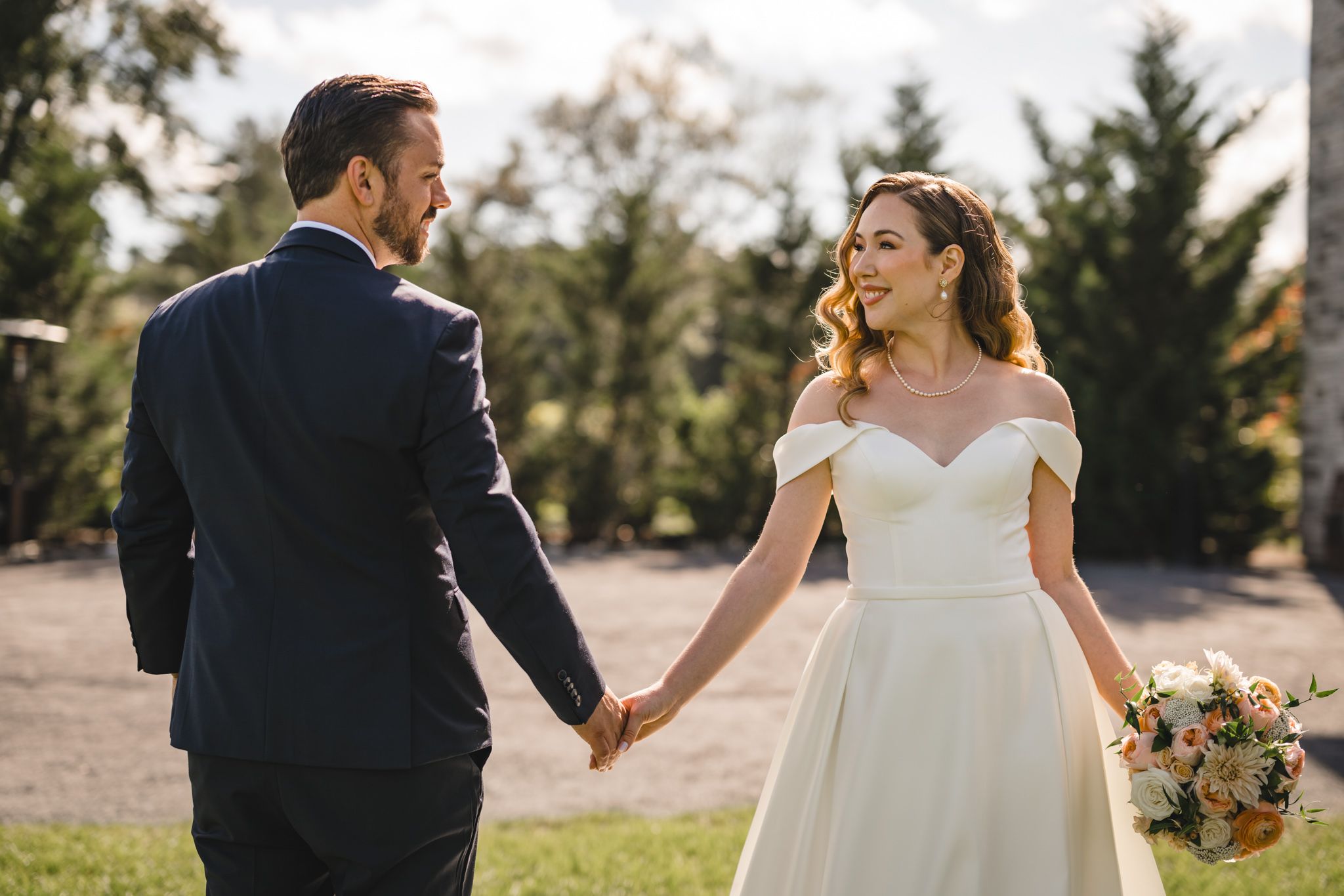 A couple holds hands while walking in formal wedding attire on a sunny day.