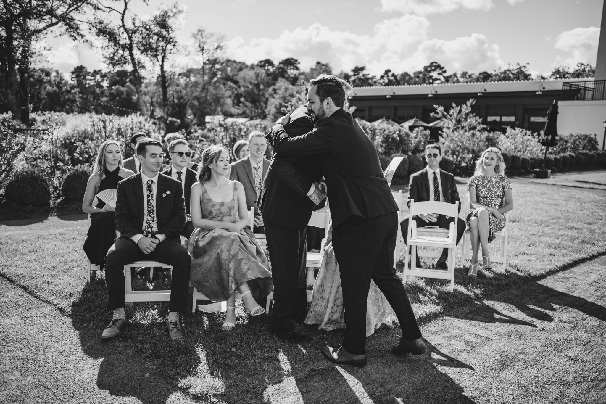 Wedding guests gather outdoors for a ceremony in black and white photography.