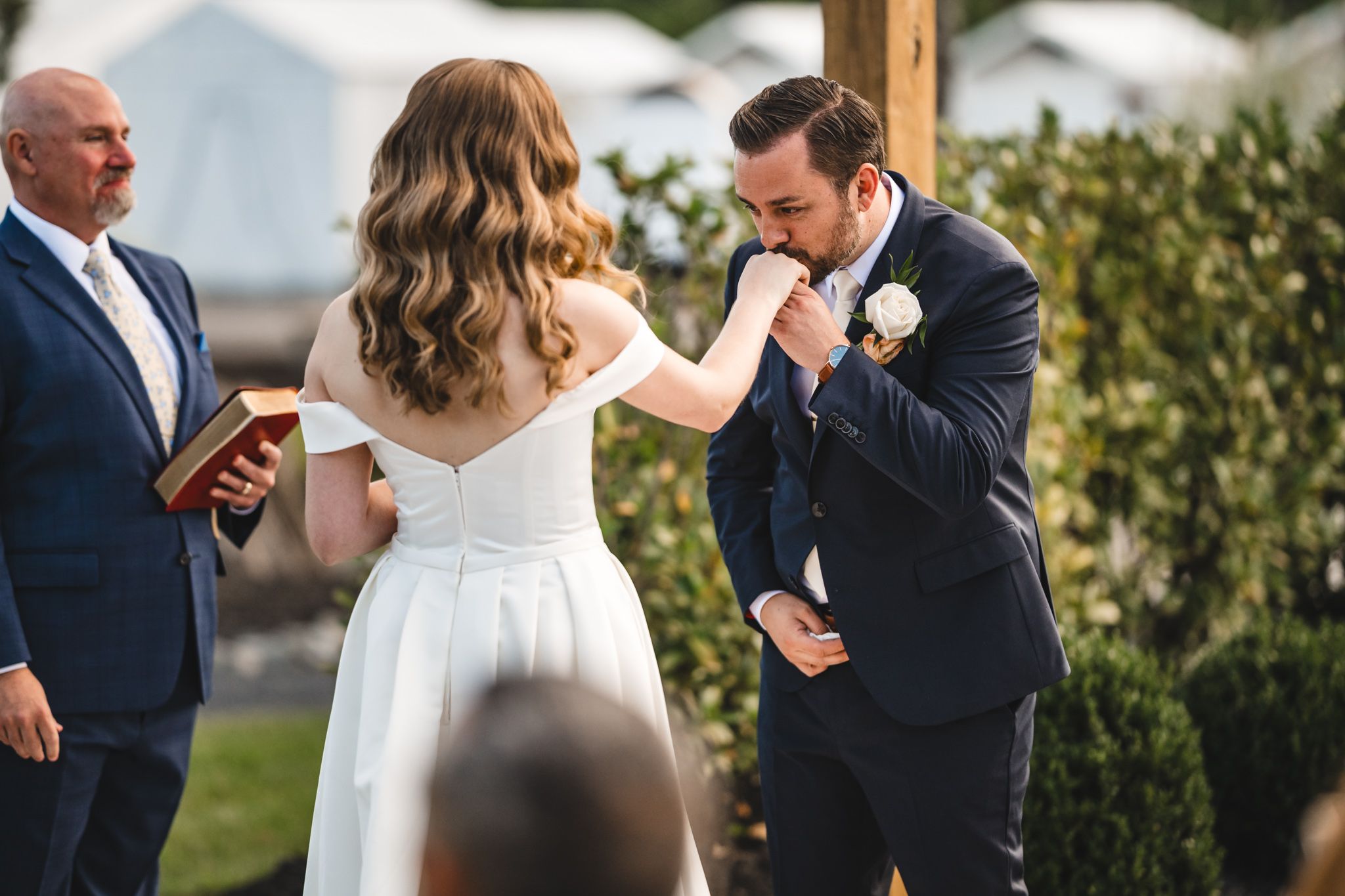 Wedding ceremony taking place under a wooden pergola in an outdoor garden setting.