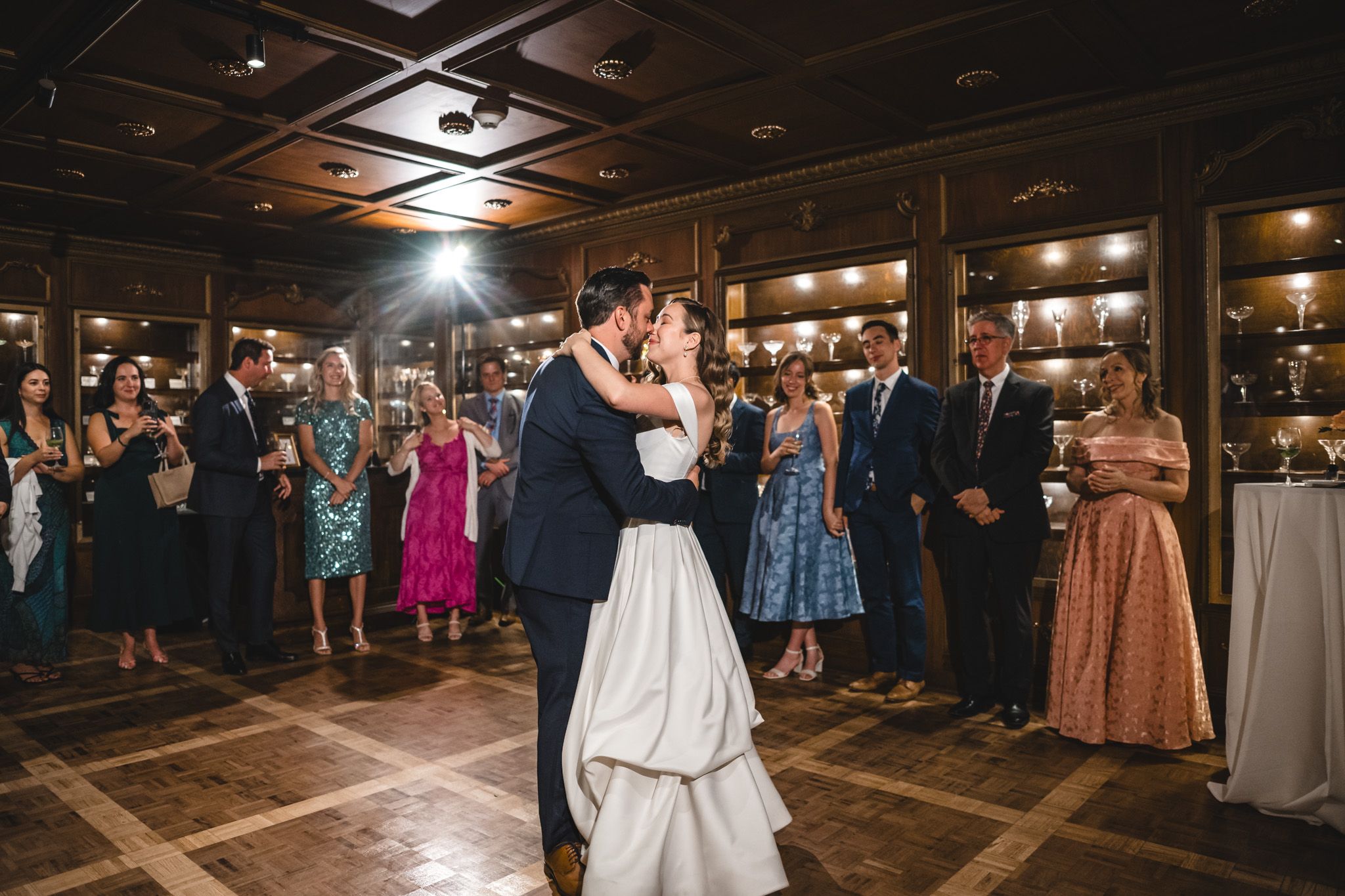 Wedding guests dance and celebrate on a rustic wooden dance floor under warm lighting at an indoor reception venue.