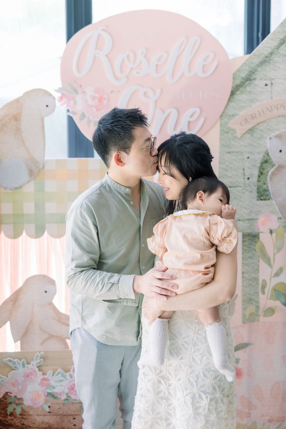 Family embraces warmly against soft pink wall in matching gray and blush outfits during portrait session.