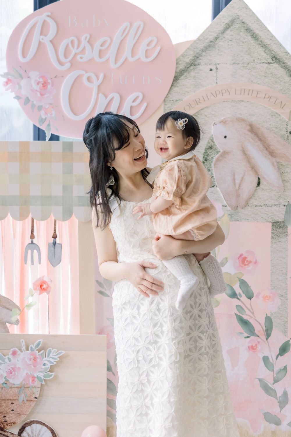Sweet mother and baby in matching white lace dresses pose against pink floral backdrop for photo series.