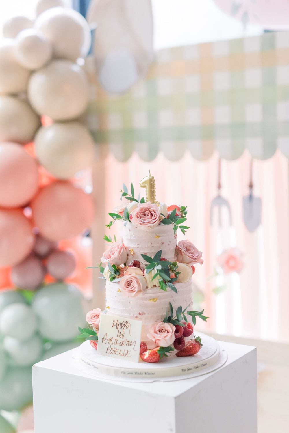 Elegant white semi-naked wedding cake decorated with pink roses and fresh strawberries on a white stand with balloon backdrop.