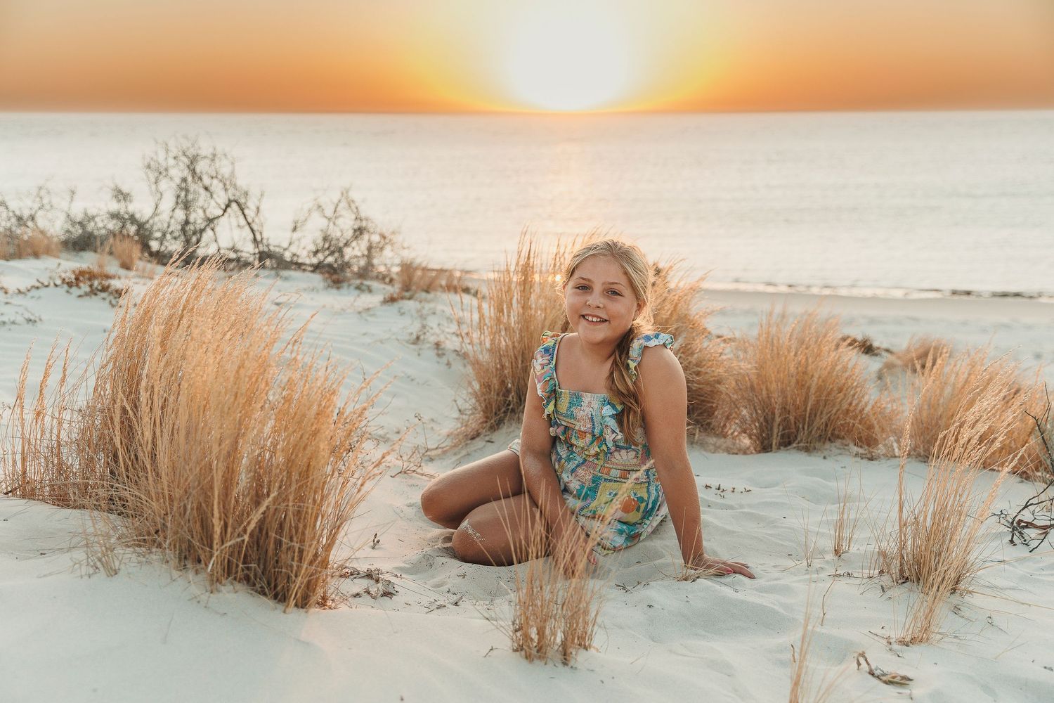 Person sitting on sandy beach at sunset surrounded by golden grass beside the ocean.